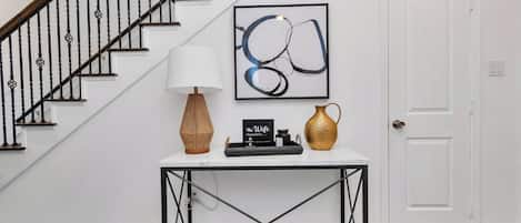 A staircase with black balusters, a white door, a table with a lamp, a vase, and a framed abstract painting.
