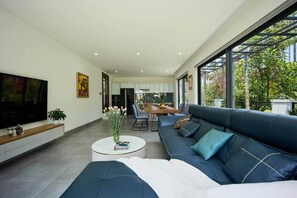A modern living room with a large TV, a sofa, a coffee table, and a dining area in the background.