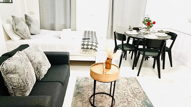 A modern living room with a black sofa, a round wooden coffee table, and a dining area with a table set for two. - One Bedroom Flat D Kentish Town (London)