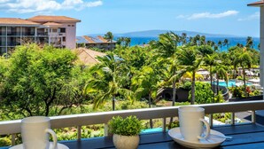 Un balcón con dos tazas y platillos blancos, una pequeña planta en maceta y vistas a un complejo turístico con palmeras y una piscina.