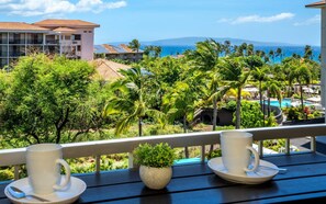 A balcony with two white cups and saucers, a small potted plant, and a view of a resort with palm trees and a pool. - Pacific Shores B-409 - Beautifully Renovated Ocean View, 2 BD / 2 BA, Split AC! (Kihei)