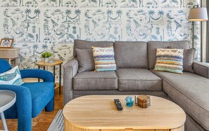 A living room with a grey sofa, blue armchair, and a round wooden coffee table with a remote control and decorative items. - Pacific Shores B-409 - Beautifully Renovated Ocean View, 2 BD / 2 BA, Split AC! (Kihei)