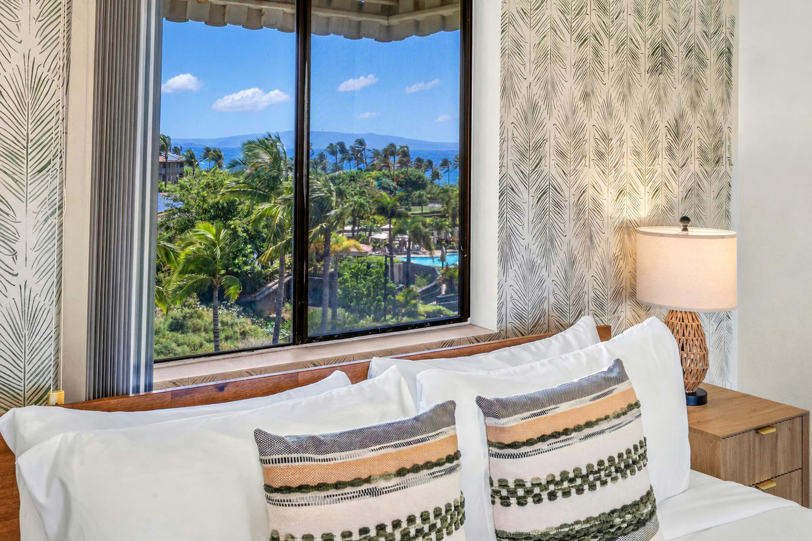 Quarto de hotel com uma grande janela que oferece vista para uma paisagem tropical, incluindo palmeiras e uma piscina.