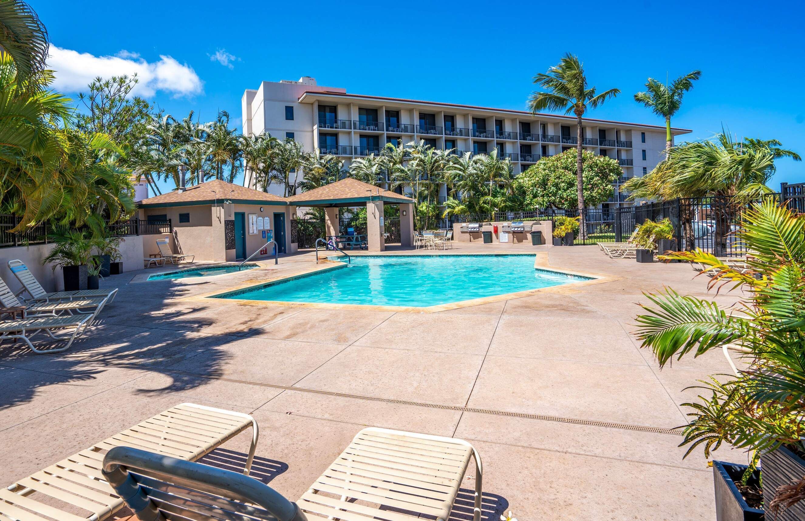 A hotel pool area with lounge chairs, a covered seating area, and a building with balconies in the background.