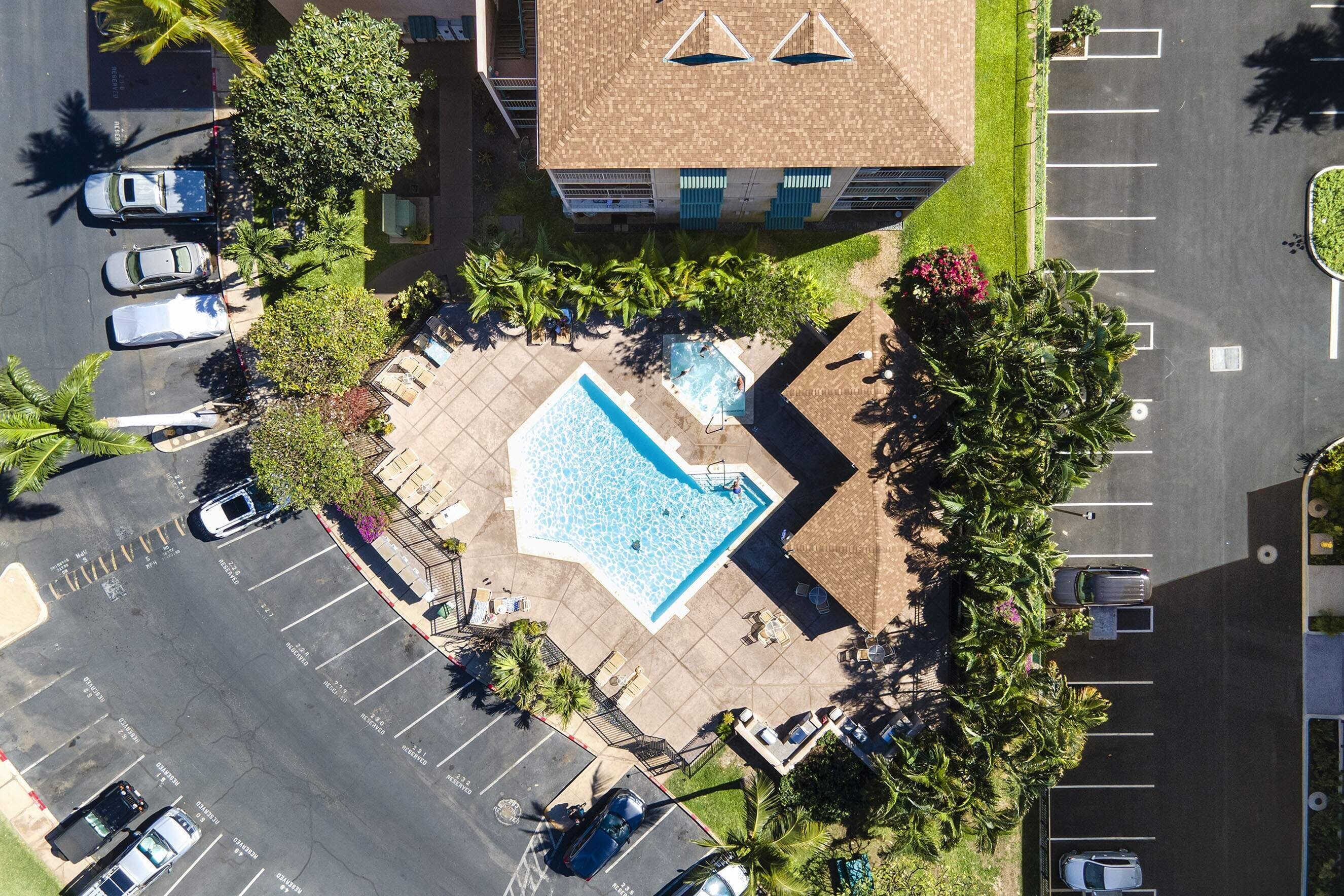 An aerial view of a parking lot with a swimming pool, surrounded by palm trees and a building with a sloped roof.