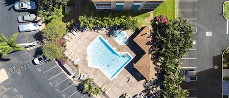 An aerial view of a parking lot with a swimming pool, surrounded by palm trees and a building with a sloped roof.