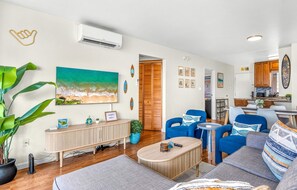 A modern living room with a large painting, a potted plant, and a wooden cabinet. - Pacific Shores B-409 - Beautifully Renovated Ocean View, 2 BD / 2 BA, Split AC! (Kihei)
