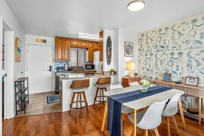 A modern kitchen with wooden cabinets, a central island, and a dining area with a blue tablecloth. - Pacific Shores B-409 - Beautifully Renovated Ocean View, 2 BD / 2 BA, Split AC! (Kihei)