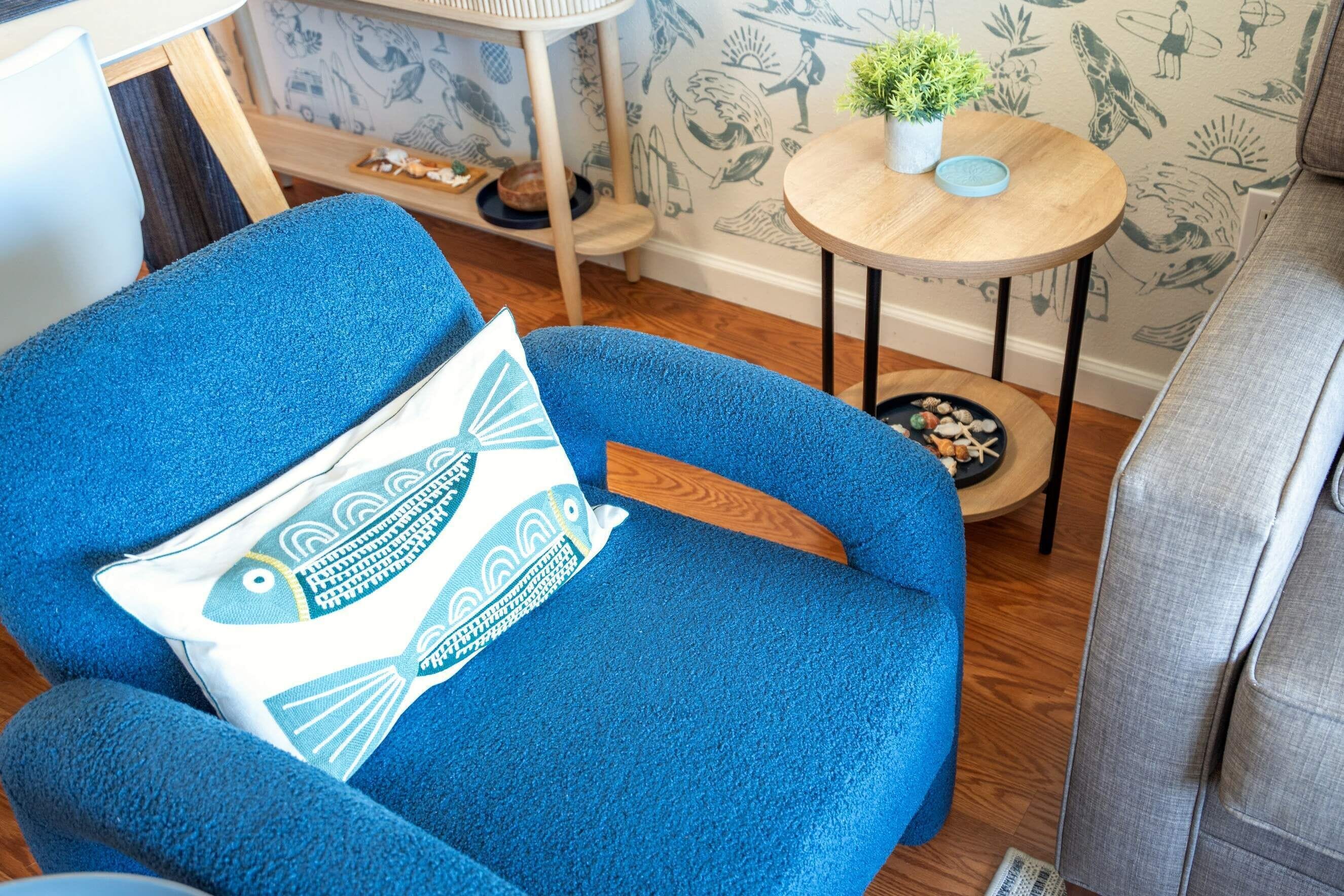 A blue armchair with a fish-patterned pillow, a small round table with a plant, and a wall with surfboard illustrations.