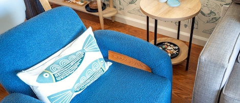 Un fauteuil bleu avec un coussin orné d’un motif de poissons, une petite table ronde avec une plante, et un mur décoré d’illustrations de planches de surf.
