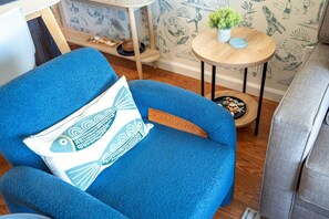 A blue armchair with a fish-patterned pillow, a small round table with a plant, and a wall with surfboard illustrations. - Pacific Shores B-409 - Beautifully Renovated Ocean View, 2 BD / 2 BA, Split AC! (Kihei)