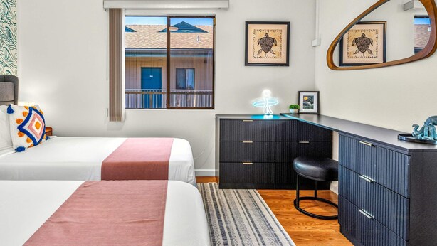 Habitación de hotel con dos camas, un escritorio, un aparador, un espejo y una ventana con vista a un edificio.