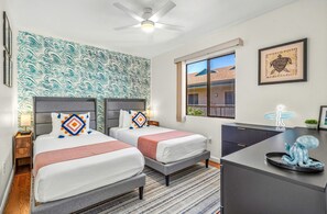 A hotel room with two beds, a desk, a ceiling fan, a window with a view, and a wall with a wave pattern. - Pacific Shores B-409 - Beautifully Renovated Ocean View, 2 BD / 2 BA, Split AC! (Kihei)
