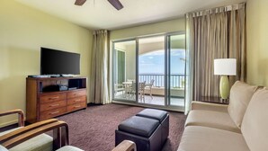 A hotel room with a flat-screen TV on a wooden cabinet, a sofa, a small table, and a balcony with a view of the sea. - Kaua‘i 2BR Ocean-View Escape – Lanai Living, Full Kitchen & Poipu Beach Nearby (Koloa)