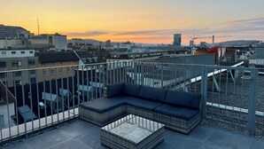 Eine Dachterrasse mit Ecksofa und kleinem Tisch mit Blick auf eine Stadtlandschaft bei Sonnenuntergang.