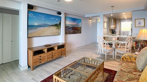 A spacious living room with a ceiling fan, a large flat-screen TV, wooden entertainment unit, and a glass coffee table. - Royal Mauian 309 - Direct Ocean Front, Renovated, Seconds to the Beach, Split AC (Kihei)
