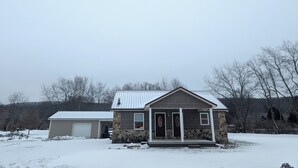 A two-story house with a stone facade and a metal roof, surrounded by snow-covered ground and leafless trees. - Golf, Relax, Repeat – Cabin Close to the Golf Course & Downtown EVL (Great Valley)