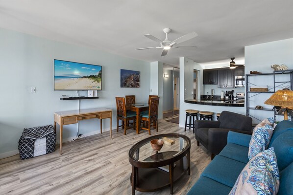 A living room with a sofa, coffee table, TV, and dining area with chairs and a table. - Kamaole Beach Royale 506 - Stunning Ocean Views, Spectacular Renovation, AC (Kihei)