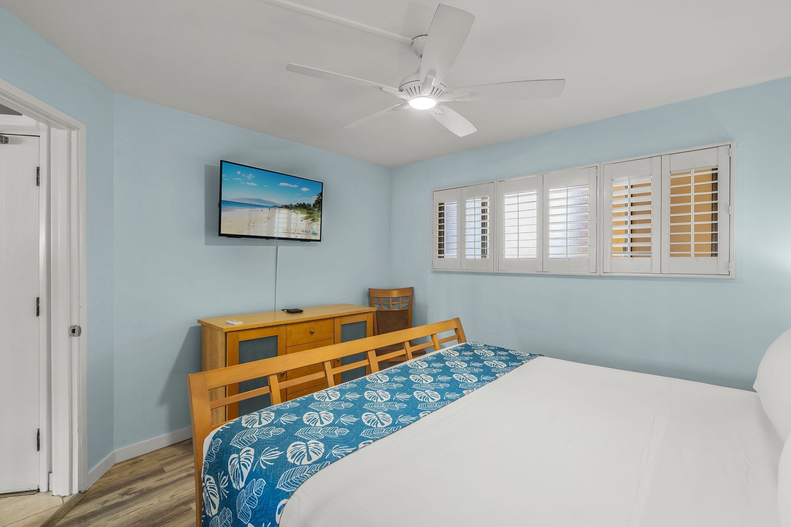 A bedroom with a bed, a desk, a chair, a ceiling fan, and a TV mounted on the wall.