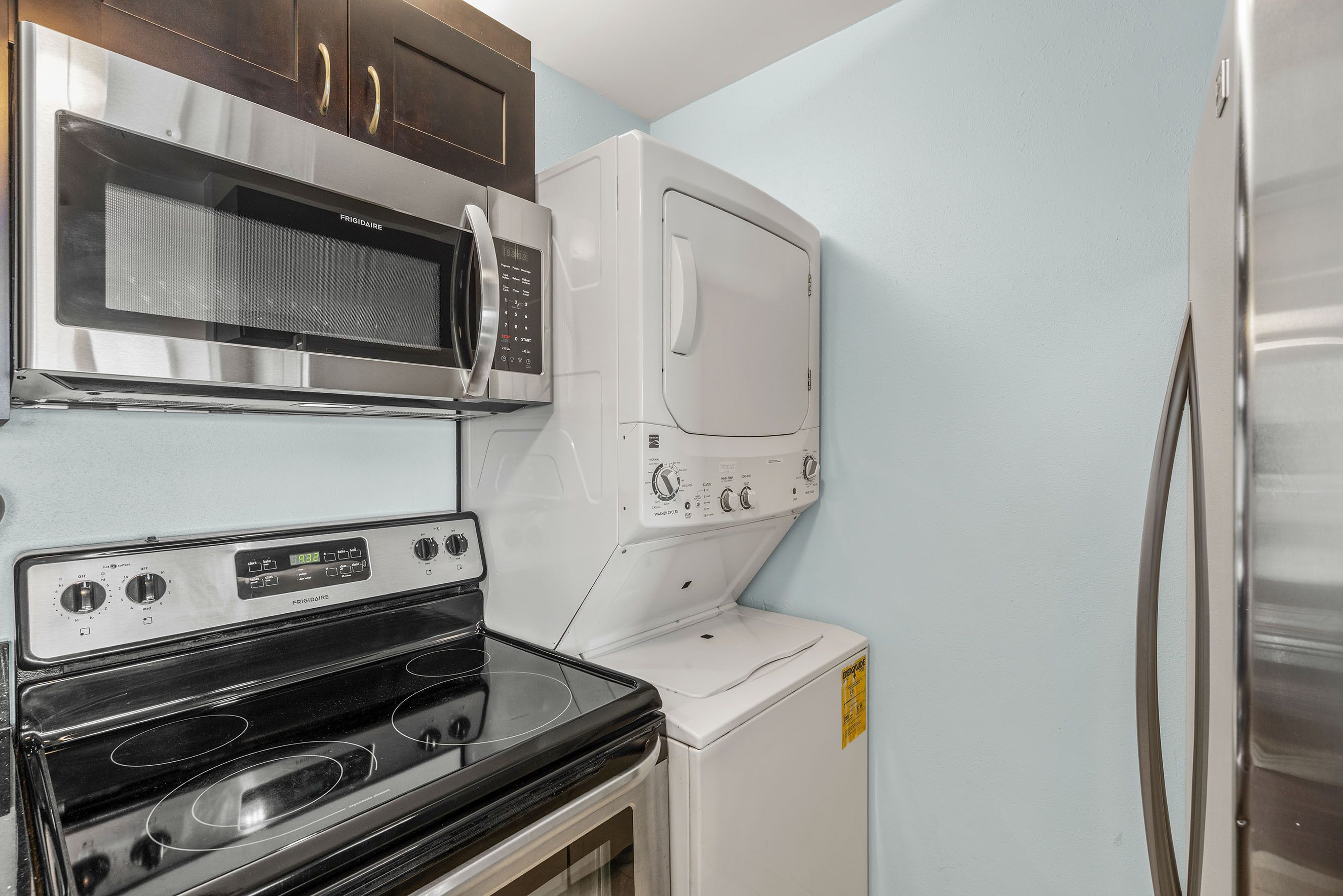 A compact kitchen with a stainless steel microwave, a built-in oven, a white washing machine, and a refrigerator.