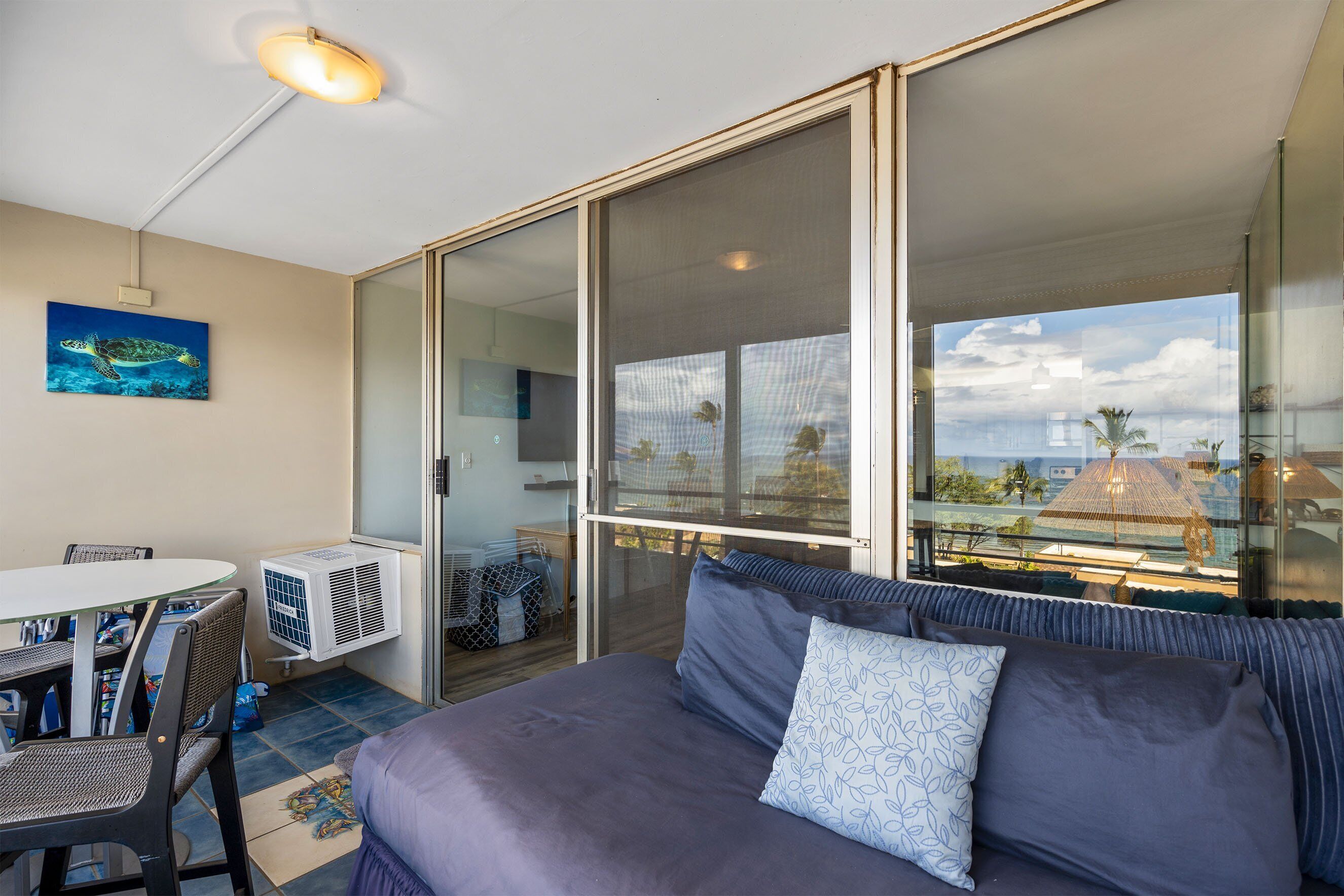 A bedroom with a bed, a sofa, a dining table, and chairs. There is a window with a view of palm trees and a building.