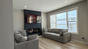 A modern living room with two grey sofas, a built-in entertainment unit, and a large window overlooking a body of water. - 
"Where the City Ends and the Mountains Begin." (Calgary)