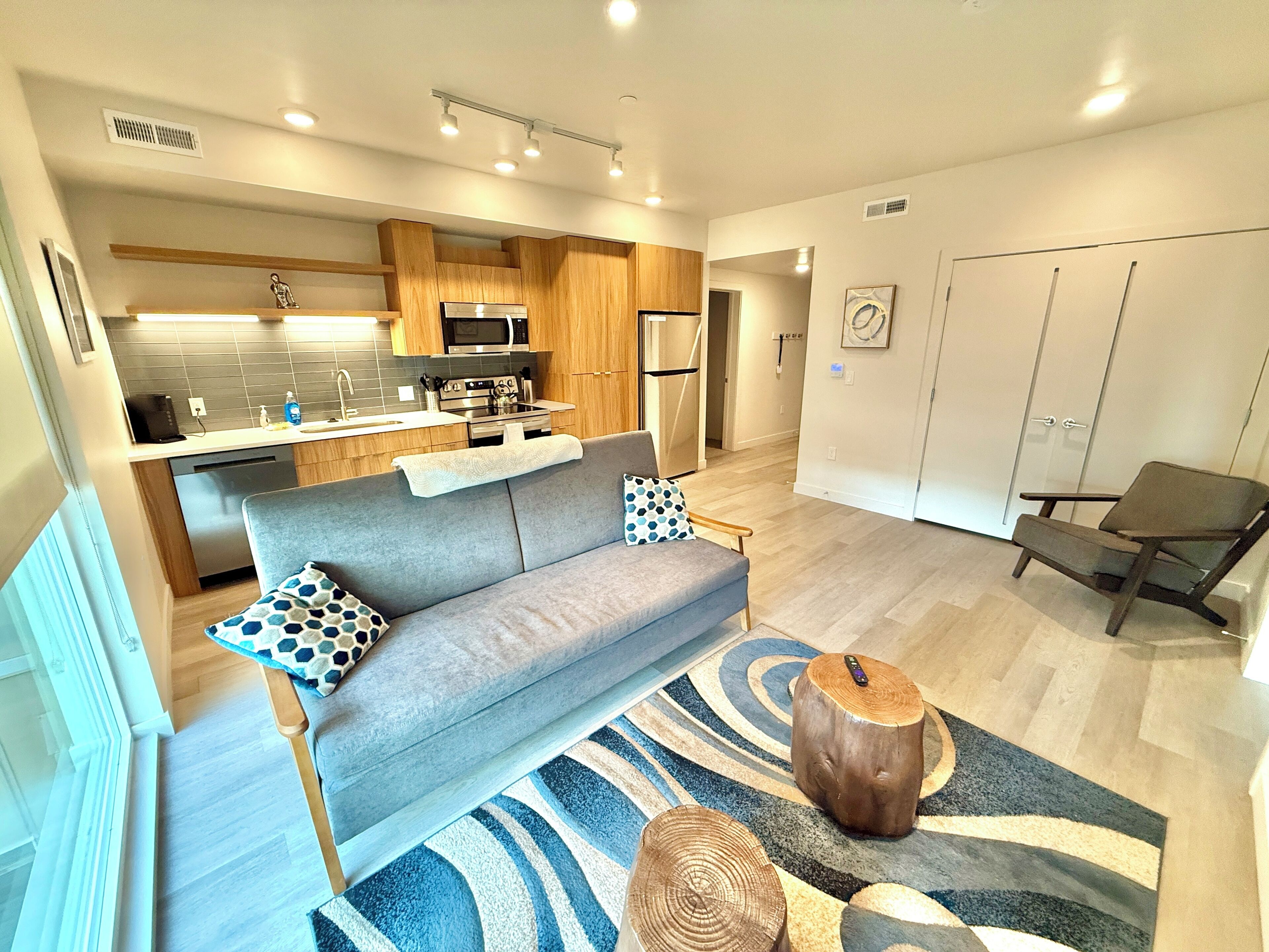 A modern apartment with a grey sofa, wooden dining table, and a kitchen area with a microwave and stove.