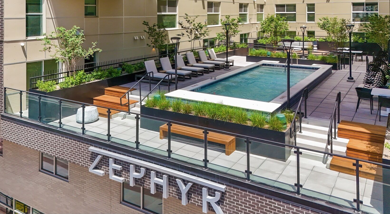 A rooftop pool area with lounge chairs and a hot tub, surrounded by greenery and a glass balustrade.