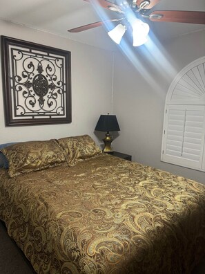A bedroom with a bed, a decorative wall piece, a ceiling fan, a lamp, and a window with shutters.