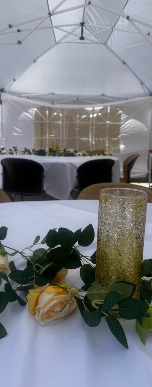 A tented event space with tables set for dining, a decorative glass centerpiece, and artificial flowers.