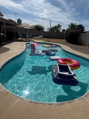 Una piscina con juguetes inflables y un patio con sillas y una parrilla.