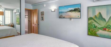 Un dormitorio con una cama, ventilador de techo, televisor montado en la pared y un cuadro.