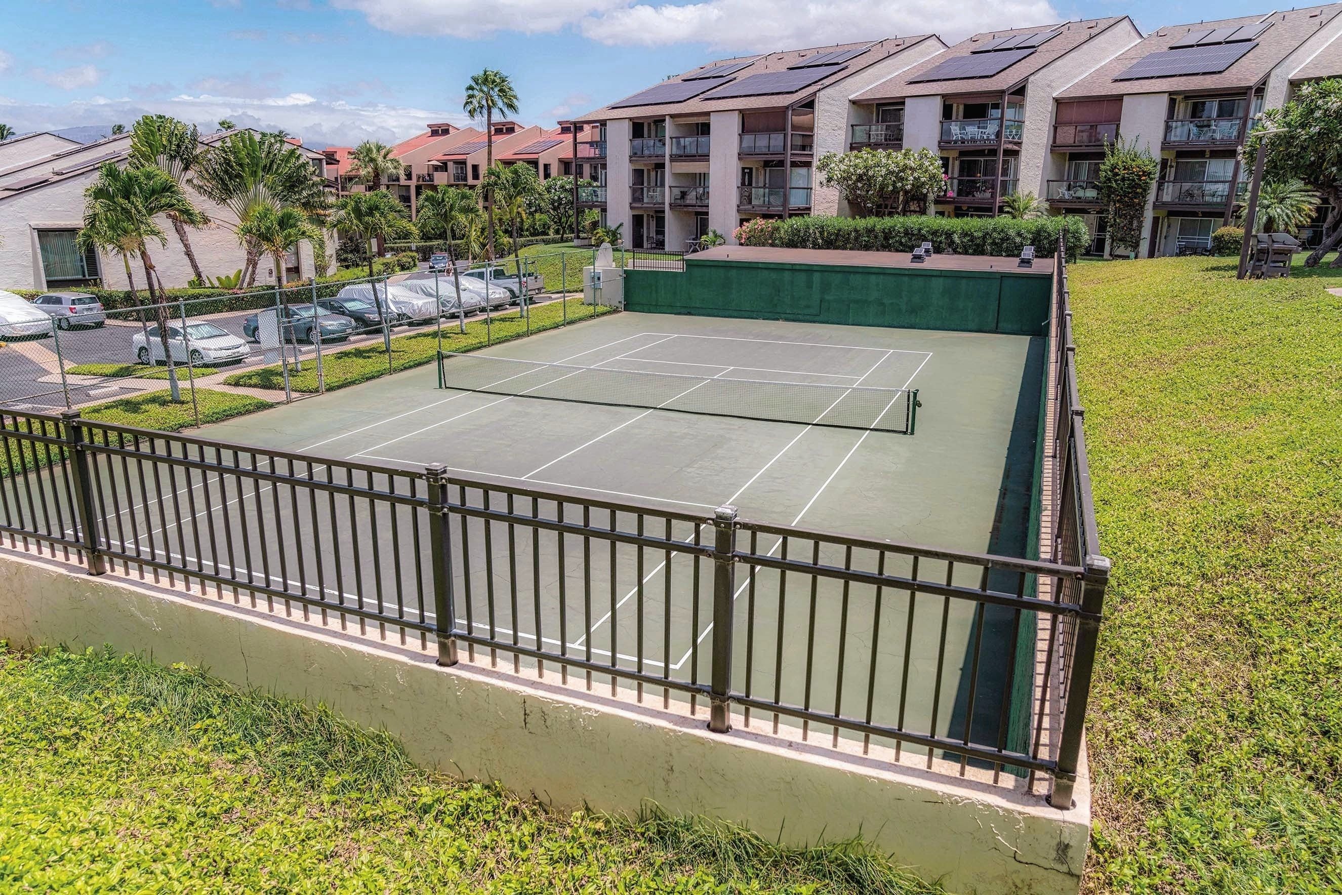 A tennis court with a surrounding fence, adjacent to a parking area and residential buildings with solar panels.