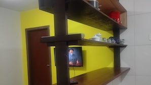 Quarto com televisão presa a uma prateleira de madeira, duas cadeiras vermelhas e uma porta ao fundo.