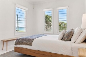 A bedroom with a large bed, two windows, a bench, and a view of the outdoors. - Miss Beachy – Stunning Beachfront Home, Pet Friendly, Amazing Views (Sapphire Beach)