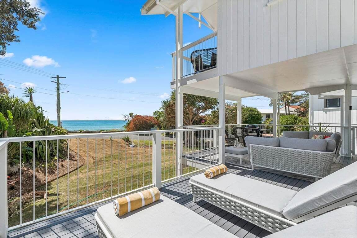 A covered outdoor seating area with a view of the ocean, white railings, and a white building in the background.