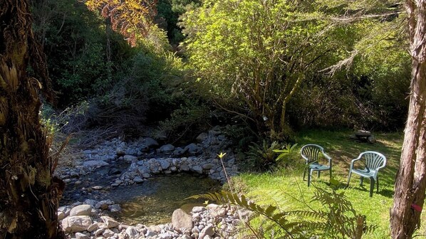 A serene garden with a small pond, surrounded by rocks and lush greenery. Two white plastic chairs are placed on the grass near the pond. - Alpacas Off Grid - Treehouse & Hut (Upper Tākaka)