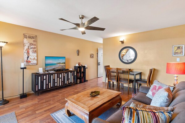 A living room with a ceiling fan, a television, a coffee table, a sofa, and a dining area with chairs and a table. - Kalama Terrace P-307 - 3rd-floor Walkup, 7 min Walk to Charley Young Beach, AC! (Kihei)