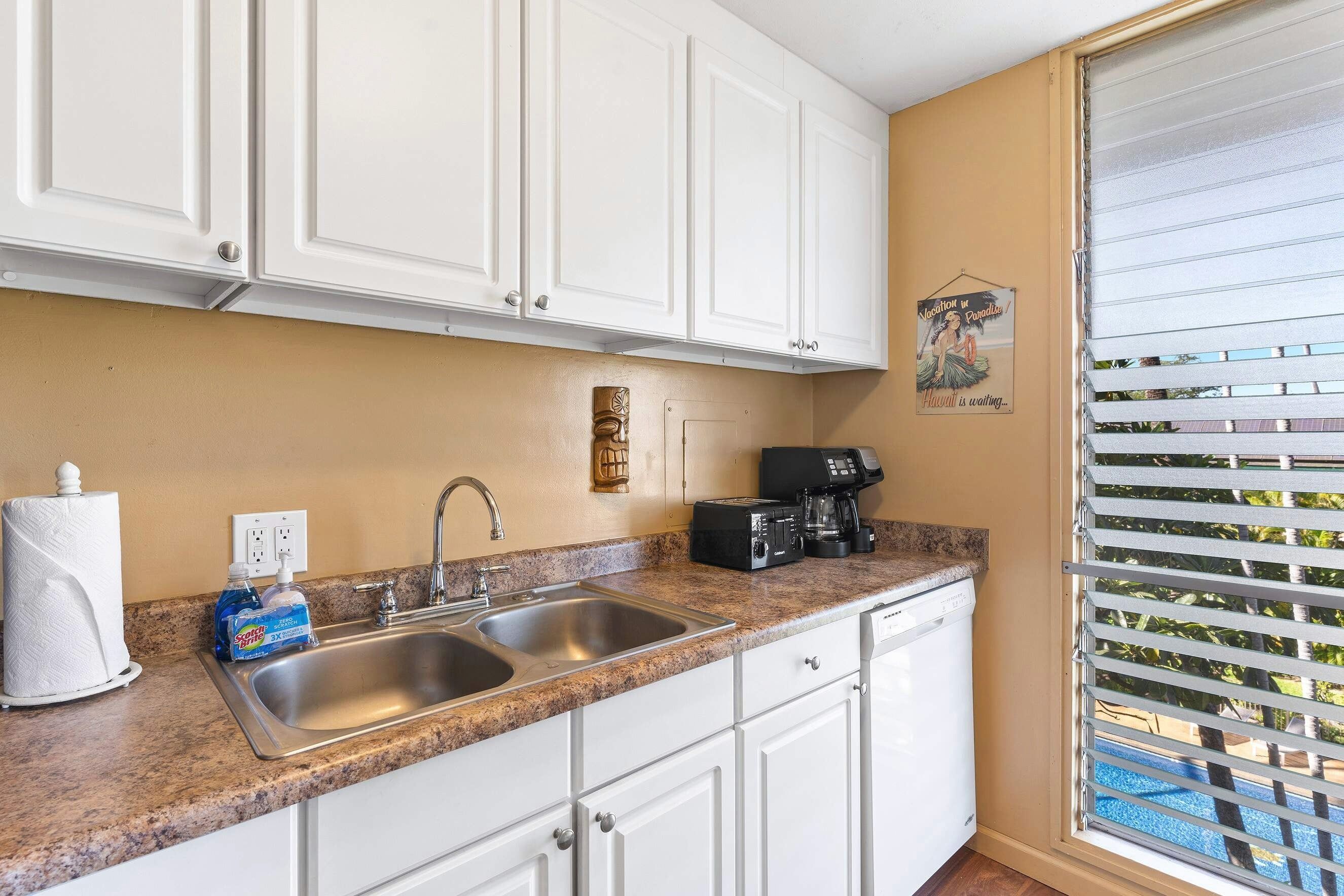 Cocina con armarios blancos, pileta doble, cafetera y vista a una piscina a través de una ventana con persianas.