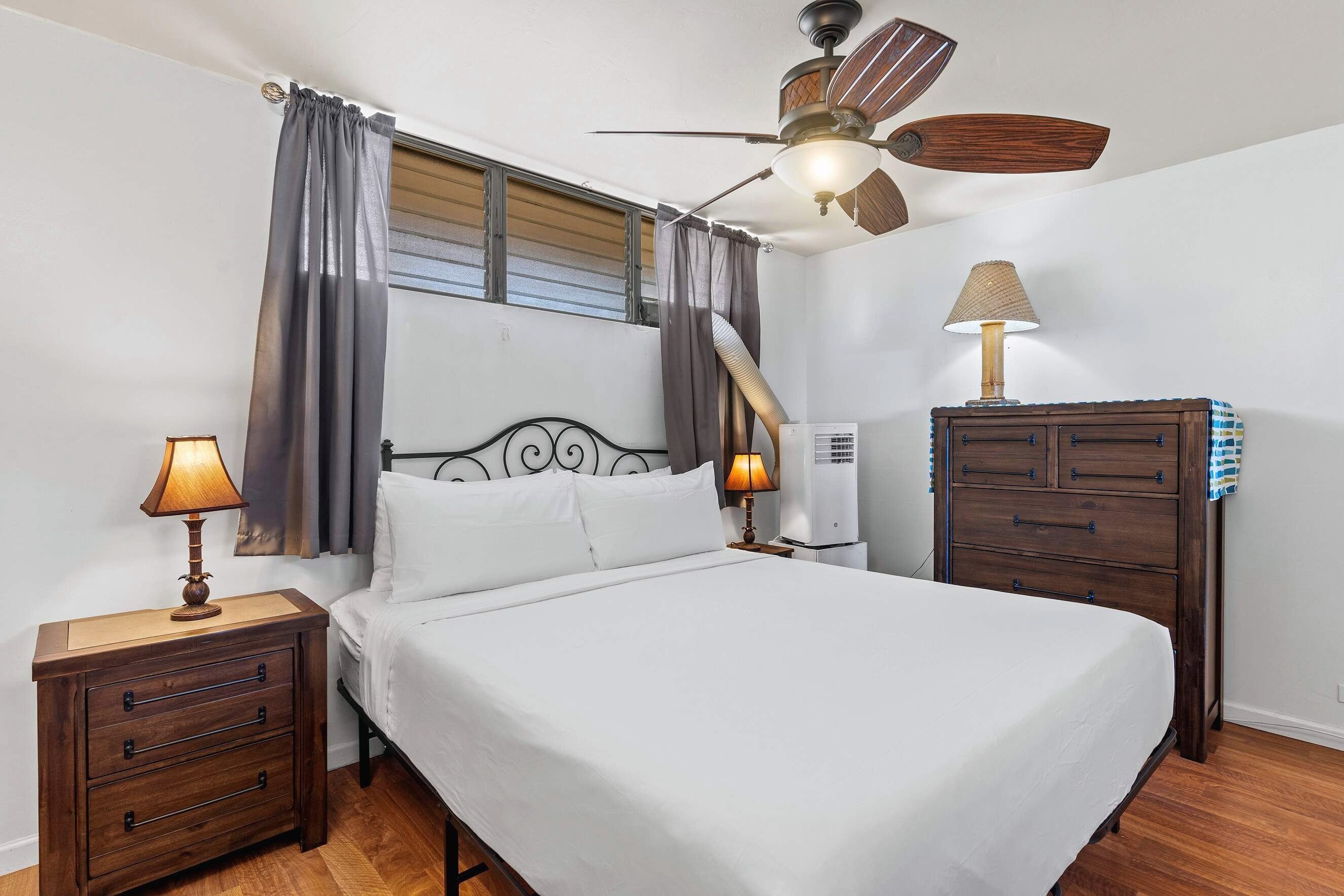 A bedroom with a bed, two bedside tables, a nightstand, a ceiling fan, an air conditioning unit, and a dresser.