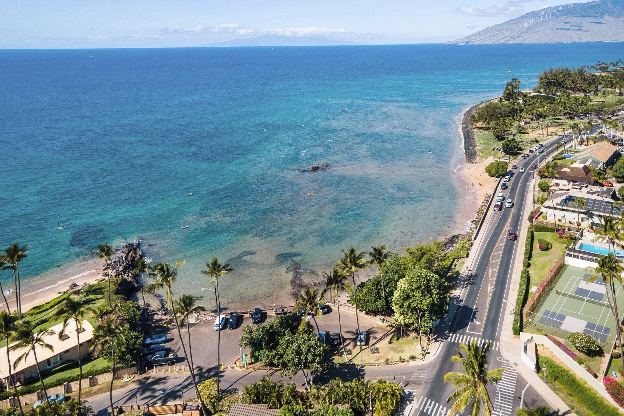 Eine Küstenstraße mit Strand auf einer Seite und Wohngebiet mit geparkten Autos und Palmen auf der anderen.