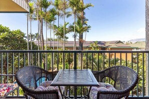 A balcony with two wicker chairs and a table, surrounded by tall palm trees and a view of buildings in the distance. - Kalama Terrace P-307 - 3rd-floor Walkup, 7 min Walk to Charley Young Beach, AC! (Kihei)