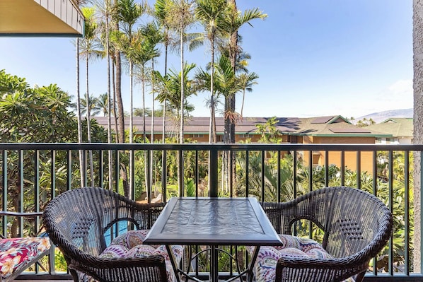 A balcony with two wicker chairs and a table, surrounded by tall palm trees and a view of buildings in the distance. - Kalama Terrace P-307 - 3rd-floor Walkup, 7 min Walk to Charley Young Beach, AC! (Kihei)