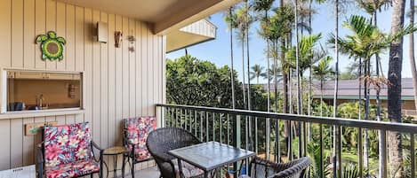 Een balkon met rieten meubels, een tafeltje en uitzicht op palmbomen en een zwembad.