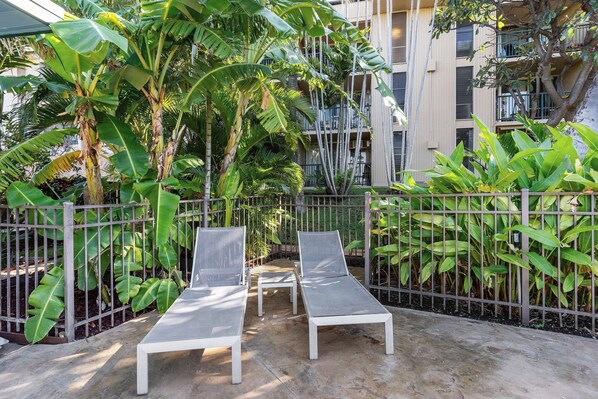 A courtyard with two lounge chairs, surrounded by a metal fence, and lush tropical plants. - Kalama Terrace P-307 - 3rd-floor Walkup, 7 min Walk to Charley Young Beach, AC! (Kihei)