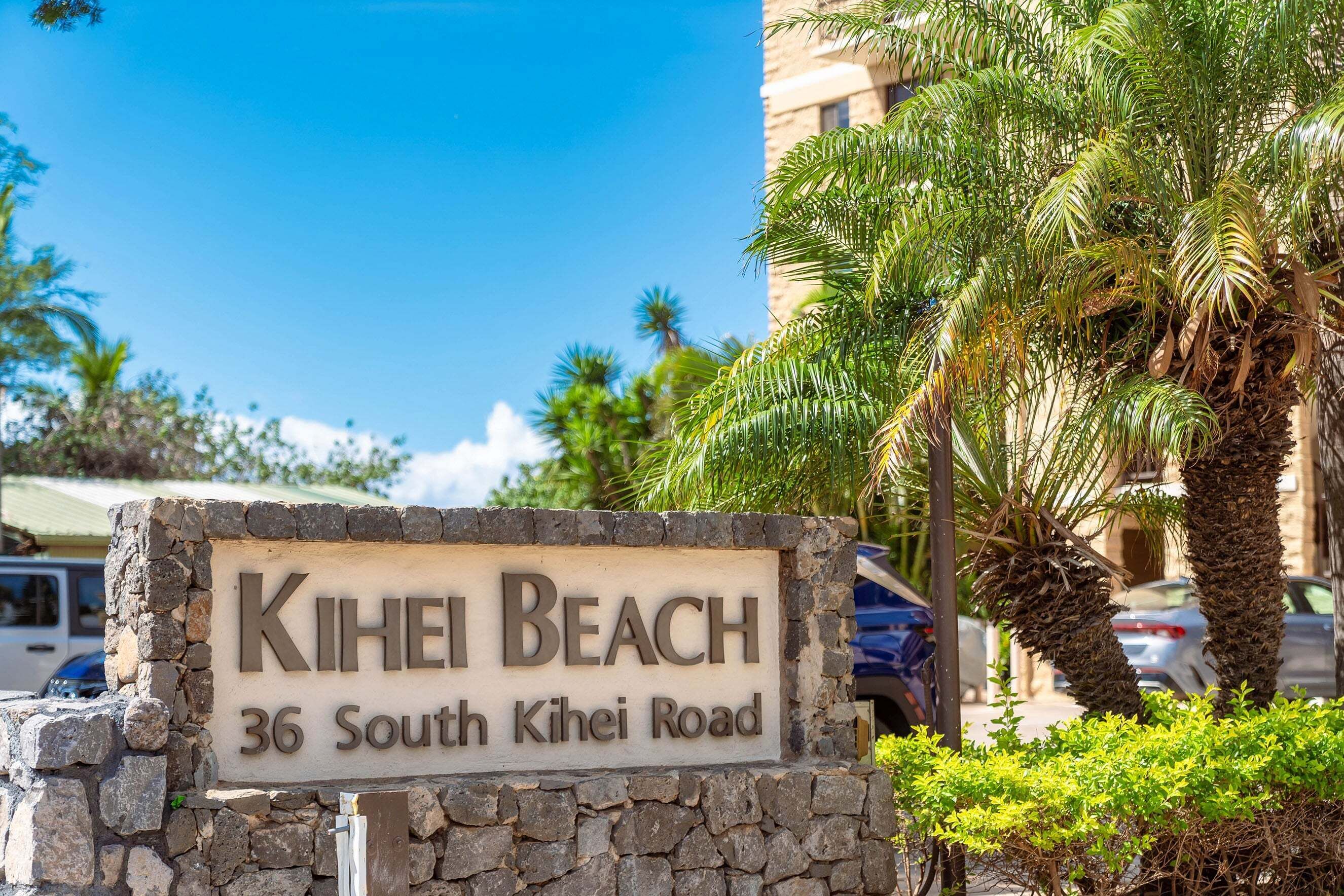 A sign for 'Kihei Beach' at 36 South Kihei Road, with a stone wall and palm trees in the background.