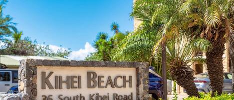 A sign for 'Kihei Beach' at 36 South Kihei Road, with a stone wall and palm trees in the background.