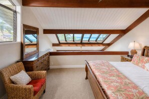 Ein Schlafzimmer mit schrägem Dach, einem Bett, einem Sessel, einer Kommode und einem Fenster mit Blick ins Freie.