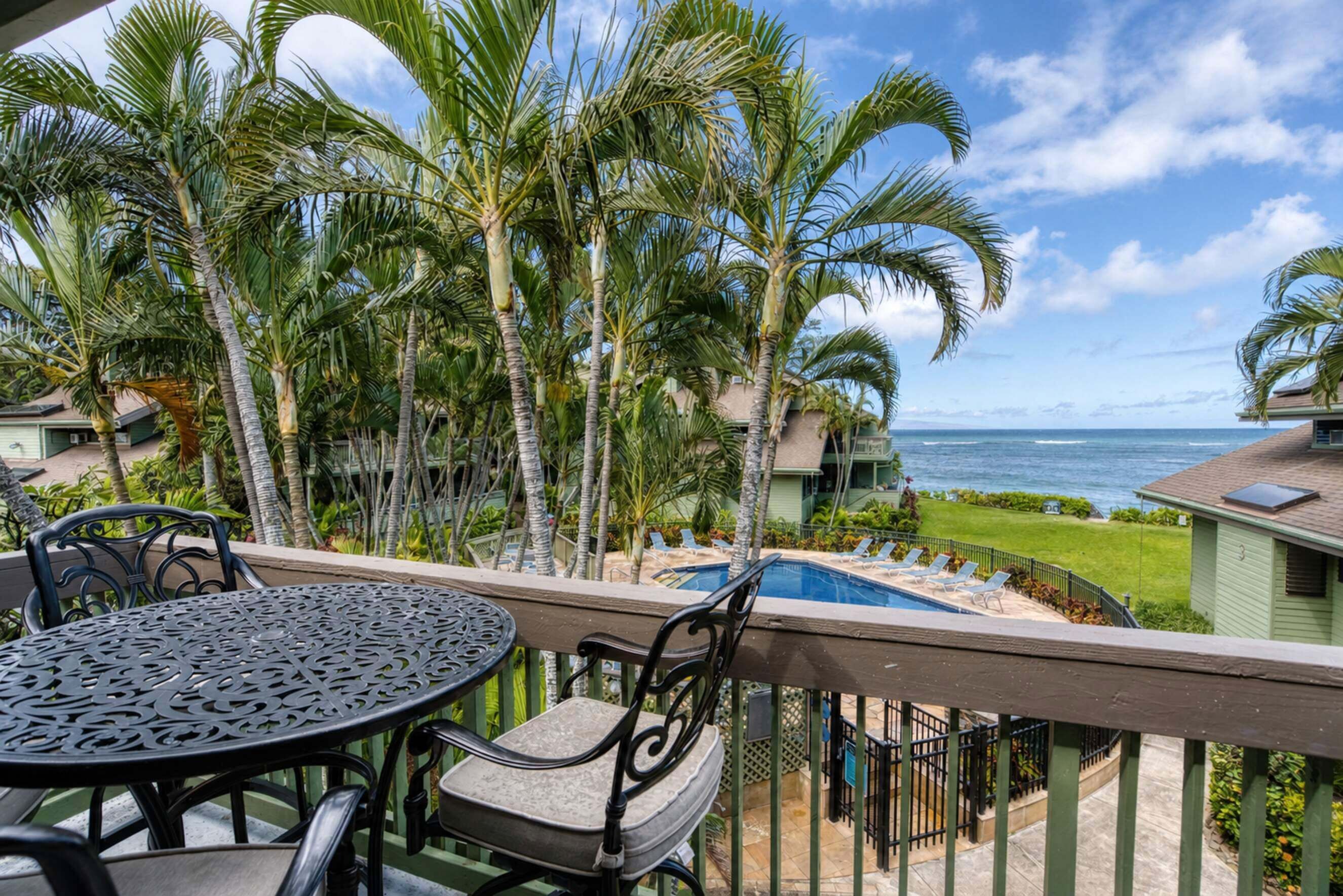 A balcony with a pool, chairs, and a table overlooking the ocean with palm trees.