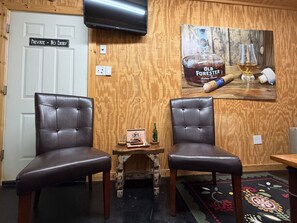 A room with two leather chairs, a small wooden table with a bottle and a framed picture, and a wall-mounted television.